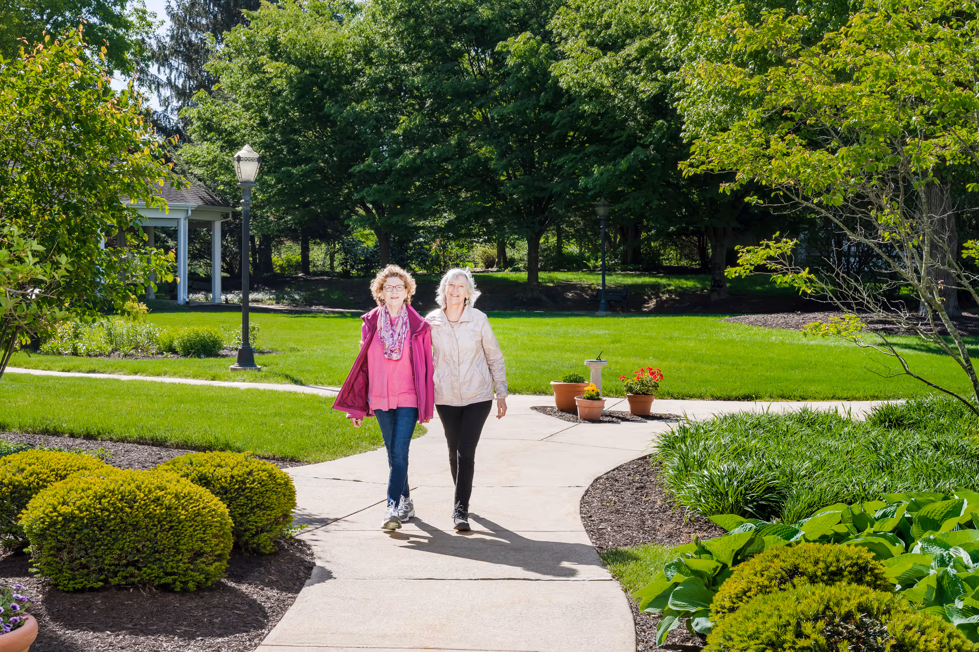 Two elderly women walking together on a paved pathway surrounded by green grass, bushes, and trees in a sunny outdoor garden area of a senior living community.