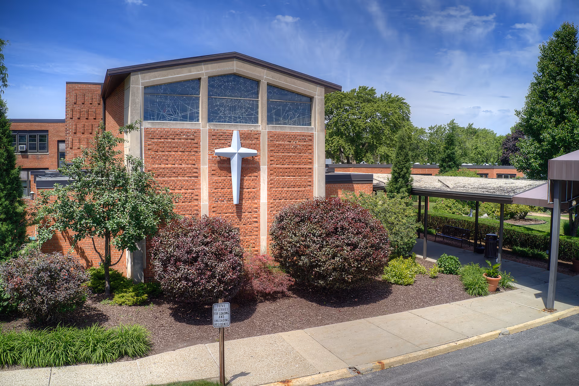Brick building exterior with a large white cross on its facade, landscaped shrubs, a sidewalk and a covered entrance.