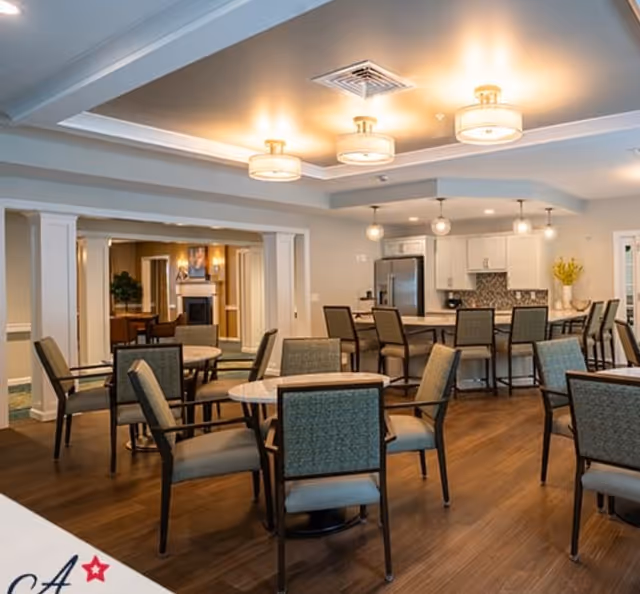 A spacious dining area in an assisted living facility featuring multiple round tables with chairs, a kitchen area with bar stools, modern lighting fixtures on the ceiling, and a warm, inviting atmosphere with wooden flooring.