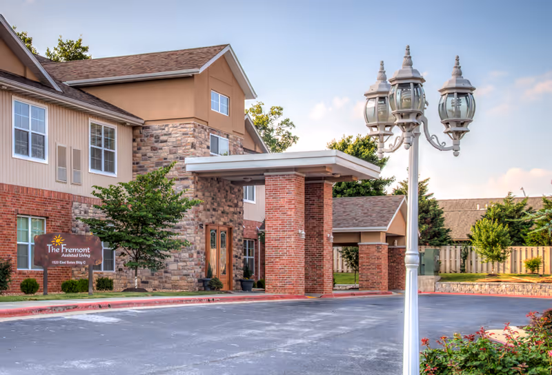 Exterior view of The Fremont Senior Living facility showing a two-story building with a combination of brick and stone facade, a covered entrance supported by brick columns, a decorative street lamp in the foreground, and landscaping with trees and bushes.