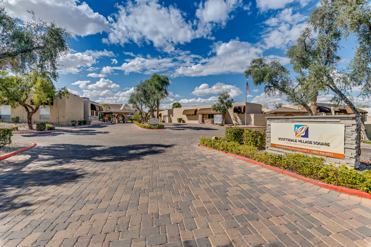 Entrance view of Scottsdale Village Square senior living community with a paved driveway, landscaped greenery, and several single-story buildings under a partly cloudy blue sky.