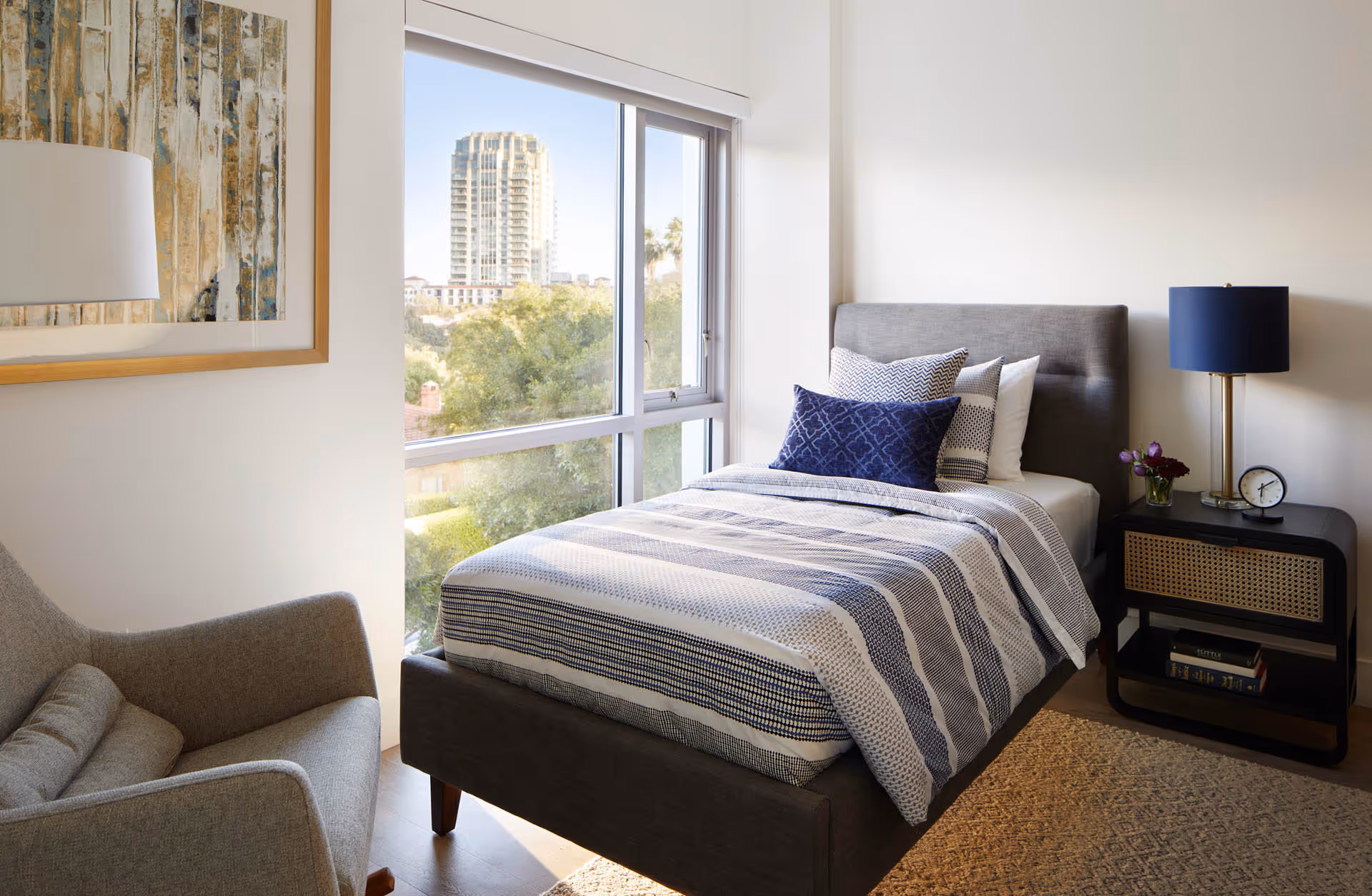 Bright single bedroom with a gray upholstered bed, patterned bedding, a nightstand and lamp, armchair, and a large window with a view of trees and a distant building.