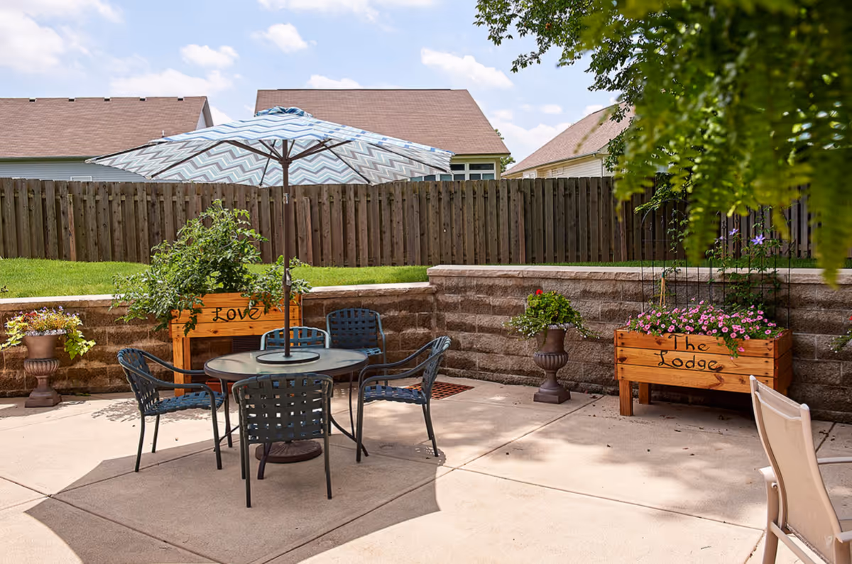 Sunny patio with a round table, umbrella, chairs, and flower planters labeled 'Love' and 'The Lodge' by a fenced lawn.