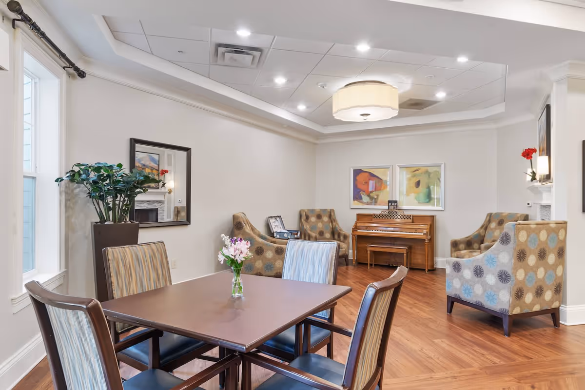 A bright and spacious common area with a square table and four chairs in the foreground, a vase with flowers on the table, patterned armchairs arranged near a wooden piano, framed artwork on the walls, a large potted plant by the window, and a ceiling light fixture.