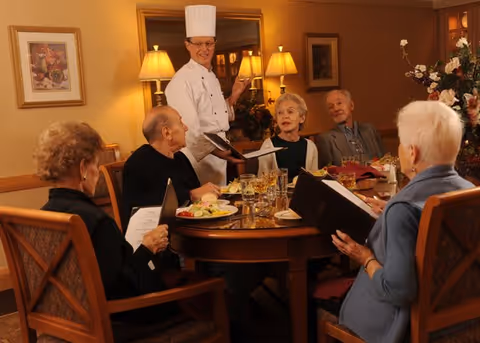 A chef in a white uniform and tall hat stands at a dining table with five elderly people seated around it. The table is set with plates of food, glasses, and menus. The room is warmly lit with lamps and decorated with framed artwork and floral arrangements.