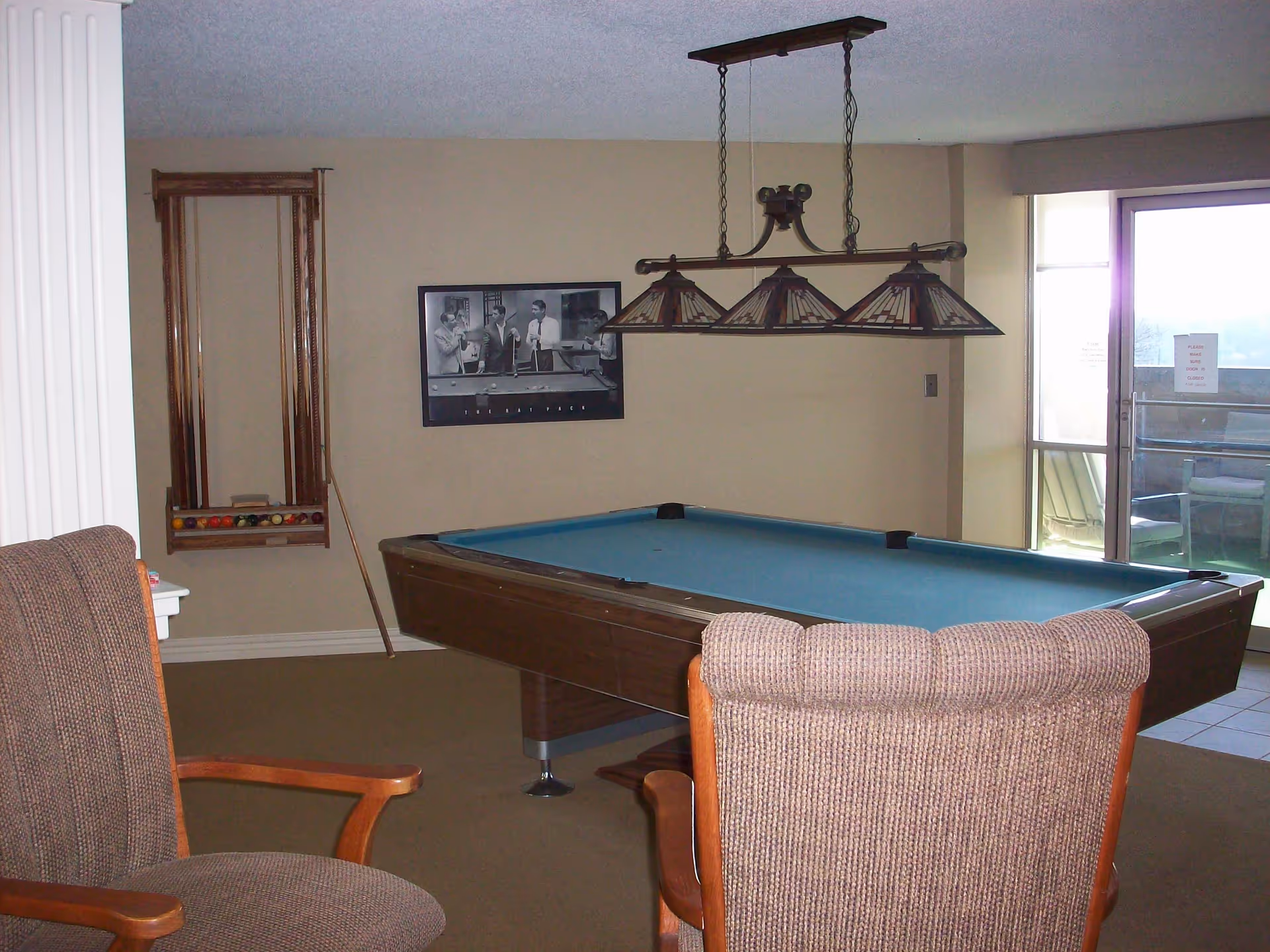 A room with a pool table covered in blue felt, two brown upholstered chairs, a wall-mounted rack holding pool cues and billiard balls, a framed black and white photo of people playing pool, and a hanging light fixture above the pool table. There is a glass door leading to an outdoor area with patio furniture.