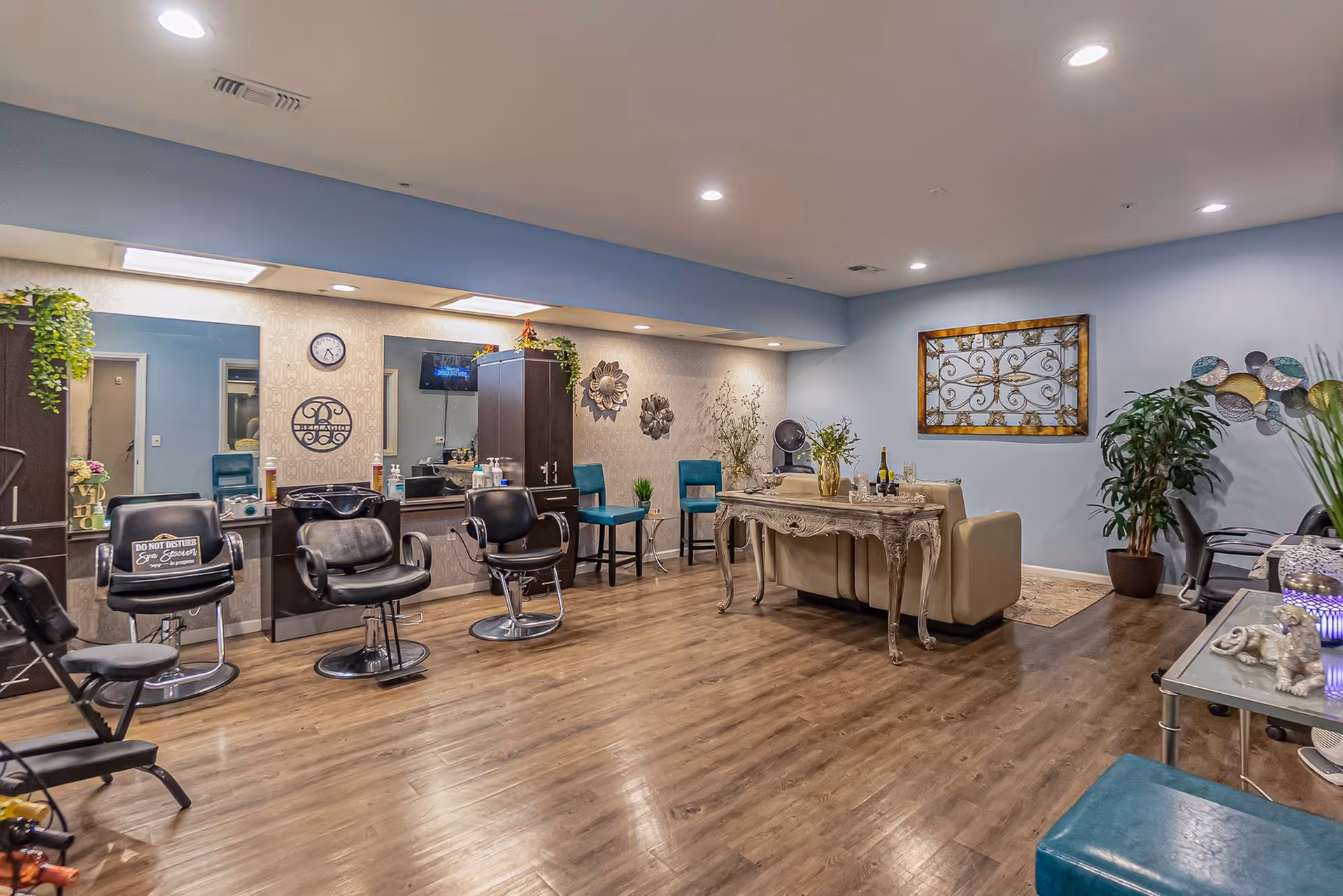 Interior view of a salon area in a senior living facility with salon chairs, mirrors, and hair washing station on the left. The room has wooden flooring, blue walls, decorative wall art, plants, and a seating area with a beige couch and a decorative table.