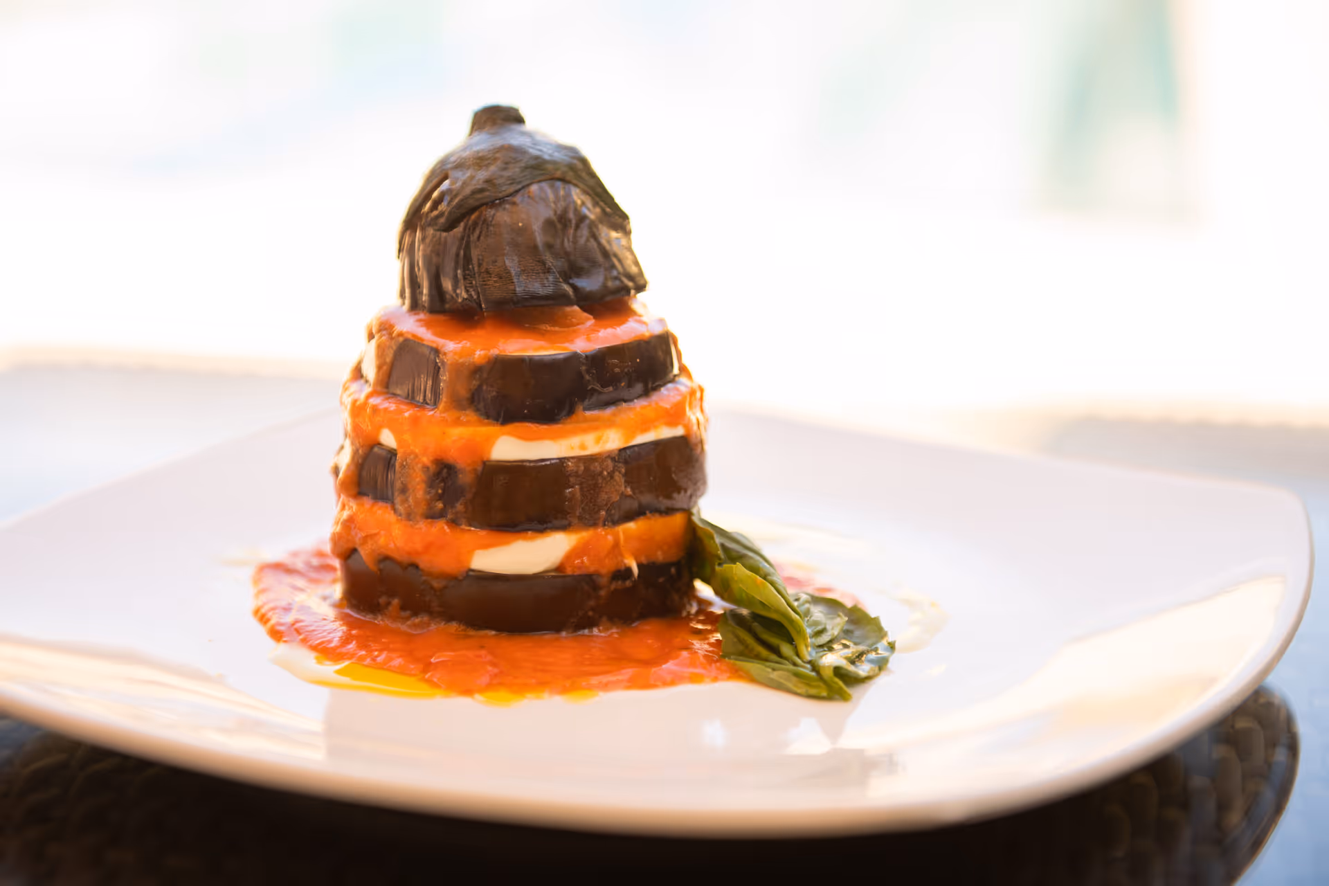 A plated dish featuring stacked slices of eggplant layered with tomato sauce and cheese, garnished with a basil leaf on a white square plate.
