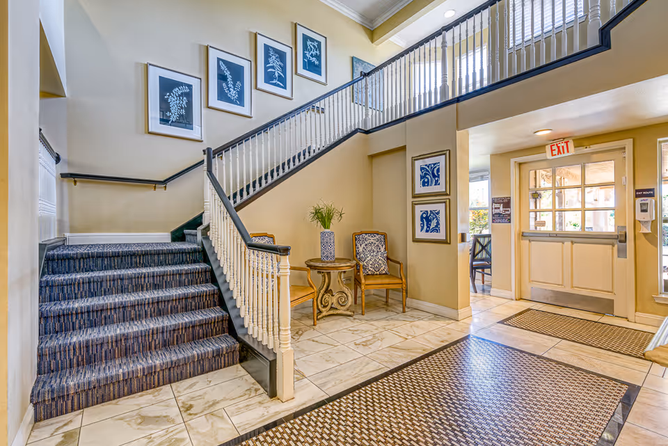 Interior view of a senior living facility entrance area featuring a carpeted staircase with white railings, beige walls adorned with framed botanical artwork, two wooden chairs with patterned cushions, a small round table with a decorative vase, tiled flooring, and a glass-paneled exit door with an exit sign above it.
