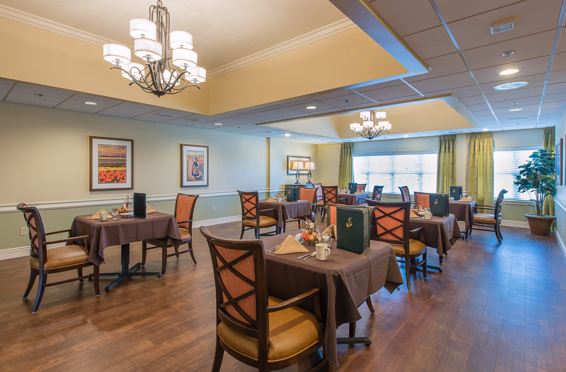 Bright dining room with several tables covered in brown tablecloths, wooden chairs, chandeliers, and large windows with green curtains.