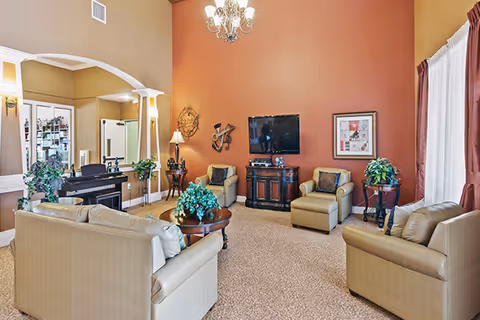 A spacious living room with beige leather sofas and armchairs arranged around a central wooden coffee table with a green plant. The walls are painted in warm tones, with one accent wall in orange. A flat-screen TV is mounted on the orange wall above a wooden cabinet. Decorative wall art and plants are placed around the room. A chandelier hangs from the ceiling, and large windows with curtains allow natural light to enter.