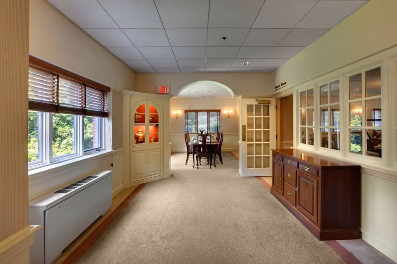 Interior view of a senior living facility hallway leading to a dining room with a wooden dining table and chairs. The hallway has beige carpet, cream-colored walls, a wooden sideboard on the right, and a window with wooden blinds on the left. There is a built-in cabinet with glass doors displaying decorative items and an exit sign above a door in the background.
