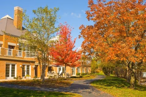 Brick senior living building exterior with a paved walkway, patio seating, and colorful autumn trees.