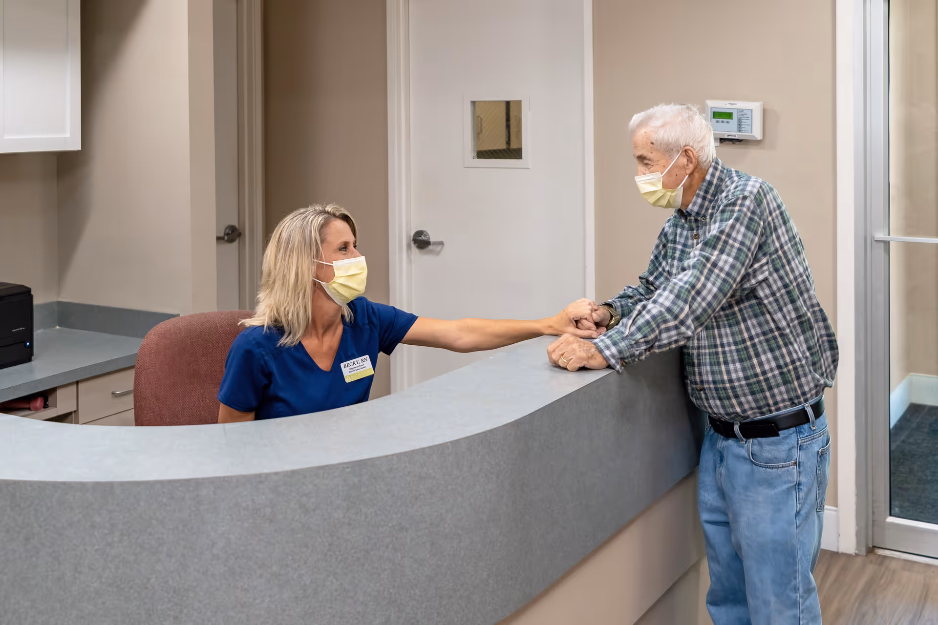 A healthcare worker wearing a blue uniform and a yellow face mask sits behind a curved reception desk, holding hands and interacting warmly with an elderly man wearing a plaid shirt, jeans, and a yellow face mask in a healthcare facility.