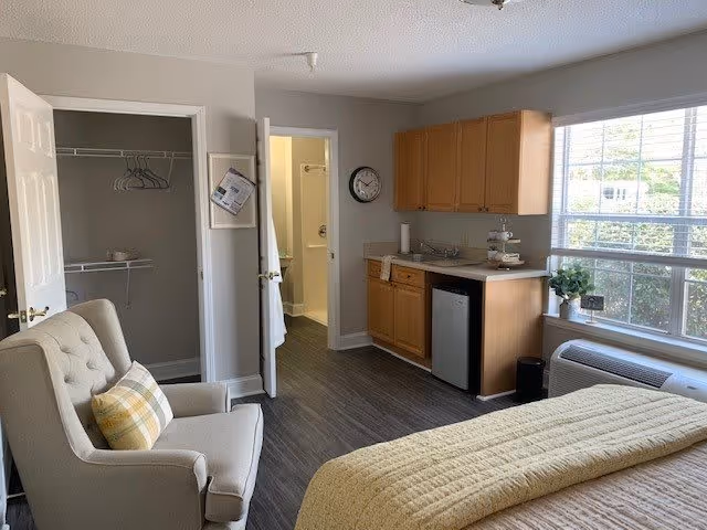 A cozy bedroom in a senior living facility with a beige armchair featuring a plaid pillow, a bed with a yellow quilt, a small kitchenette with wooden cabinets, a mini fridge, and a sink. There is a window letting in natural light, a wall clock, and an open closet and bathroom door visible.