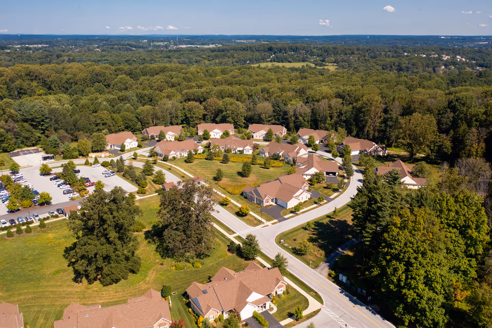 Aerial view of Kendal At Longwood senior living community showing multiple single-story residential buildings with brown roofs arranged in a circular layout surrounded by trees and greenery, with a parking lot visible on the left side.