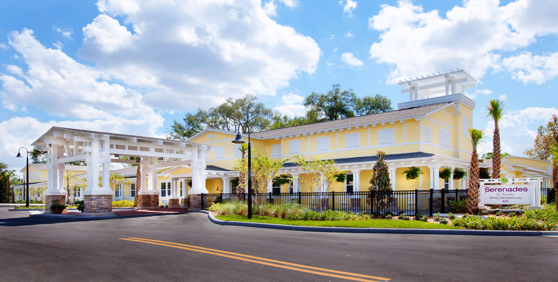 Exterior view of a yellow two-story senior living facility building with white trim and a covered entrance. The building is surrounded by landscaping including palm trees and bushes, under a partly cloudy blue sky. A sign in front reads 'Serenades by Sonata Memory Care Assisted Living 425'.