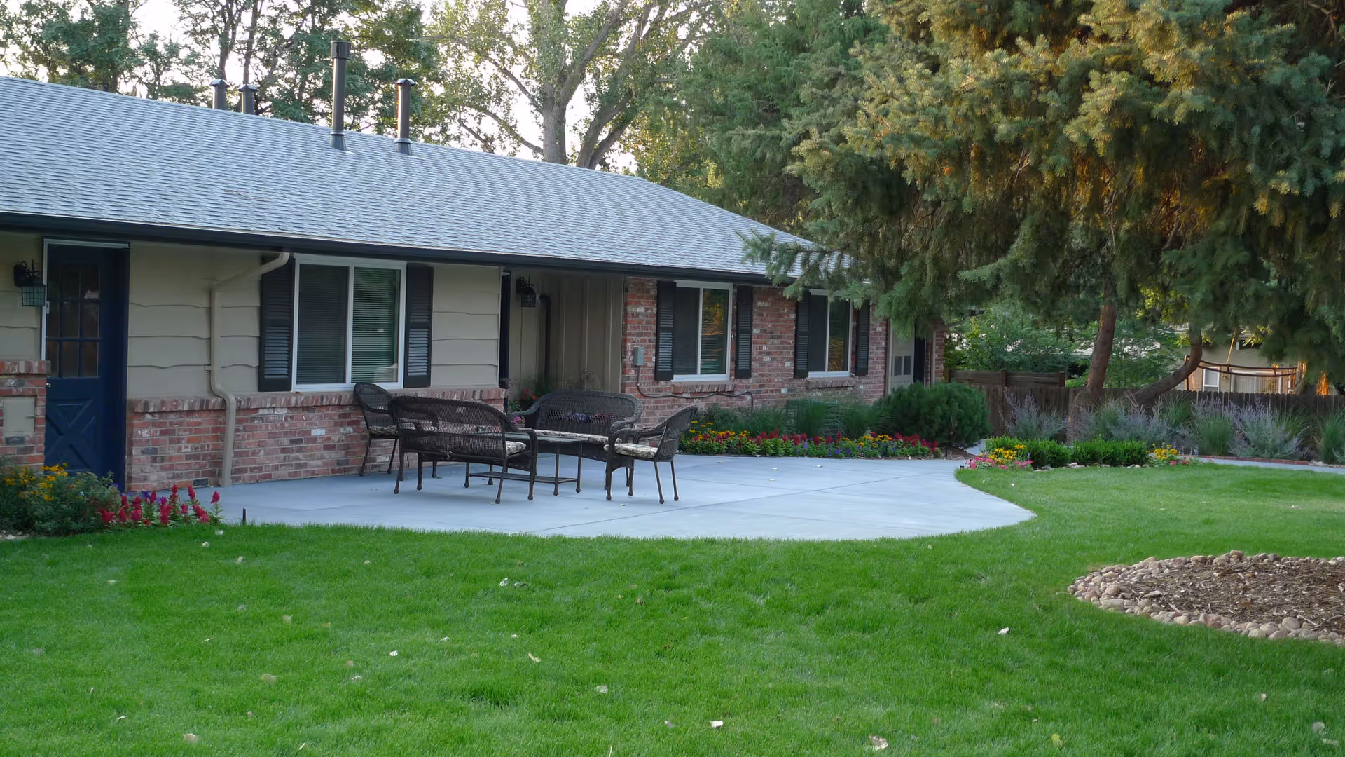 Outdoor patio area of a single-story brick and siding building with a concrete seating area furnished with metal chairs and a table. The patio is surrounded by green grass, flower beds, and trees providing shade.
