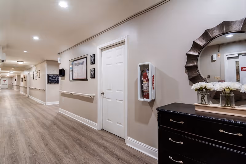 A long, well-lit hallway in a senior living facility with beige walls and wood flooring. On the right side, there is a black dresser with white flowers in glass vases and a large decorative round mirror above it. A fire extinguisher is mounted on the wall next to a white door. The hallway has handrails along the walls and framed pictures and signs, including one that says 'Welcome to Chronicles'.