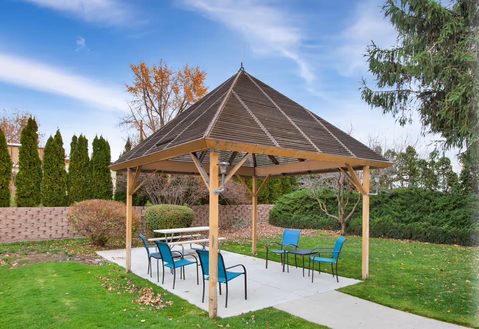 Outdoor gazebo with a wooden frame and slatted roof situated on a concrete pad surrounded by green grass and various bushes and trees. Under the gazebo are a white picnic table and several blue chairs arranged around a small black table. The sky is partly cloudy.