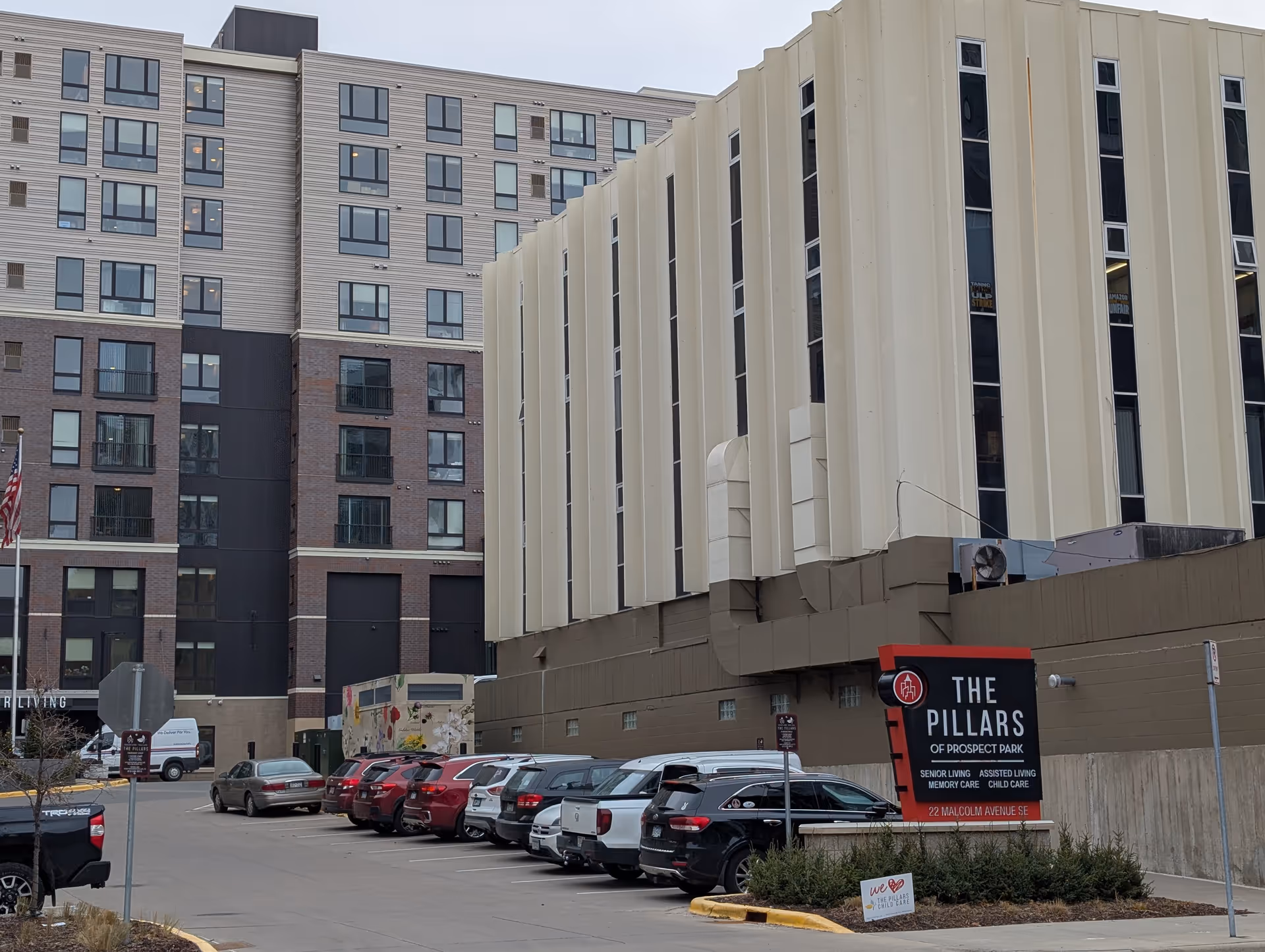 Exterior view of The Pillars of Prospect Park building, its parking lot, and a red entrance sign.