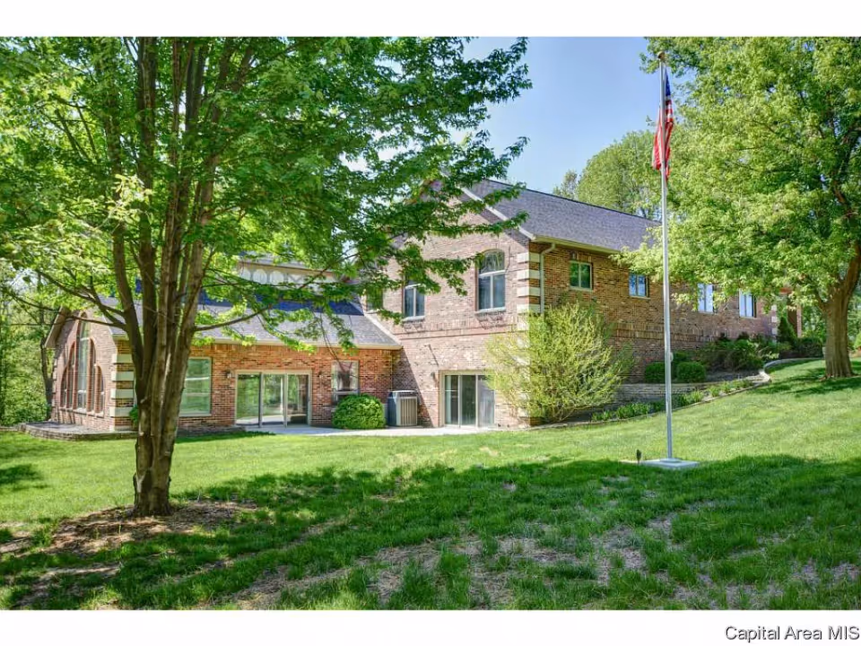 Exterior view of a brick building surrounded by green grass and trees under a clear blue sky, with an American flag on a flagpole in the foreground.