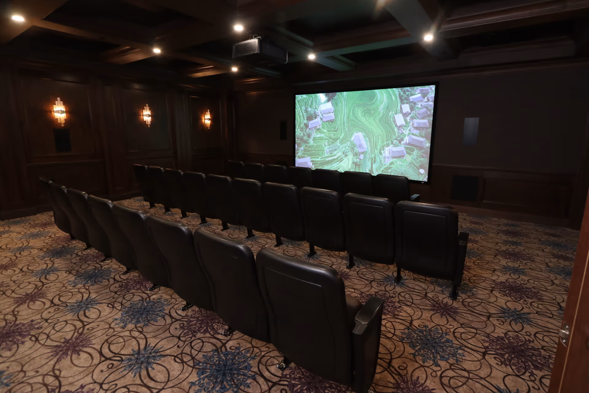 A small theater room with two rows of black leather recliner chairs facing a large screen displaying an aerial view of houses and green terrain. The room has dark wood paneling on the walls, decorative wall sconces emitting warm light, and a patterned carpet with blue and purple floral designs.