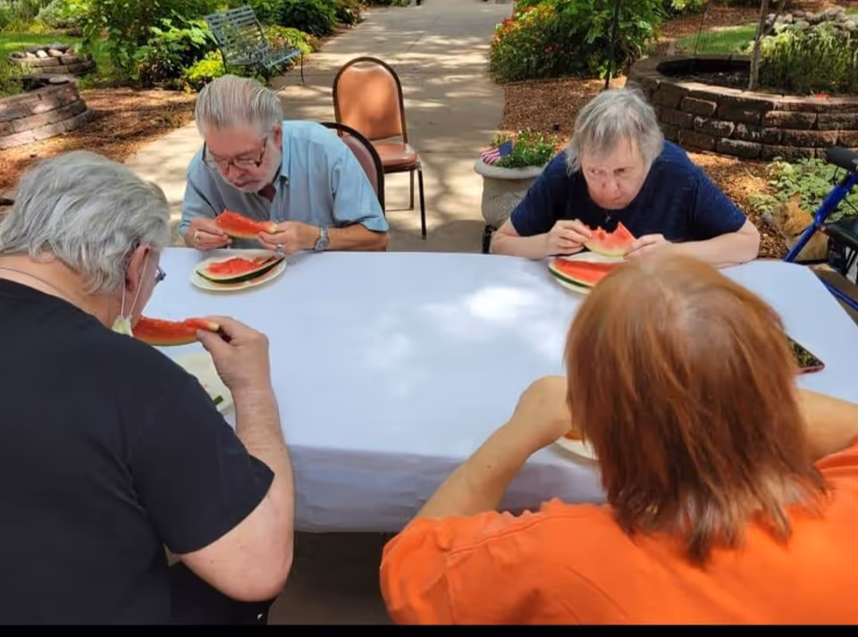 Four elderly individuals sitting around a white table outdoors, eating slices of watermelon. The setting appears to be a garden or patio area with greenery and a paved walkway in the background.