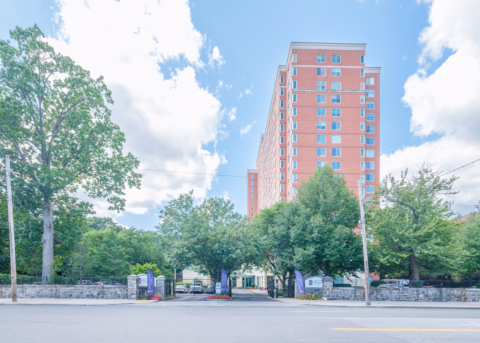 Exterior view of a tall red brick residential building behind a gated entrance with stone walls and trees on either side under a partly cloudy sky.