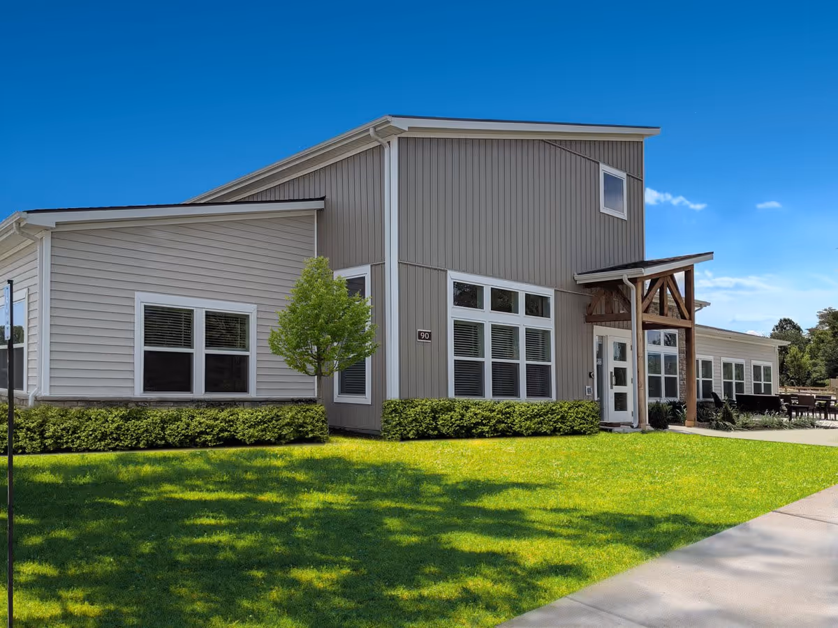 Exterior view of a modern single-story building with beige and gray siding, multiple windows, a small tree, and neatly trimmed bushes in front. The building has a covered entrance with wooden beams and a well-maintained green lawn under a clear blue sky.
