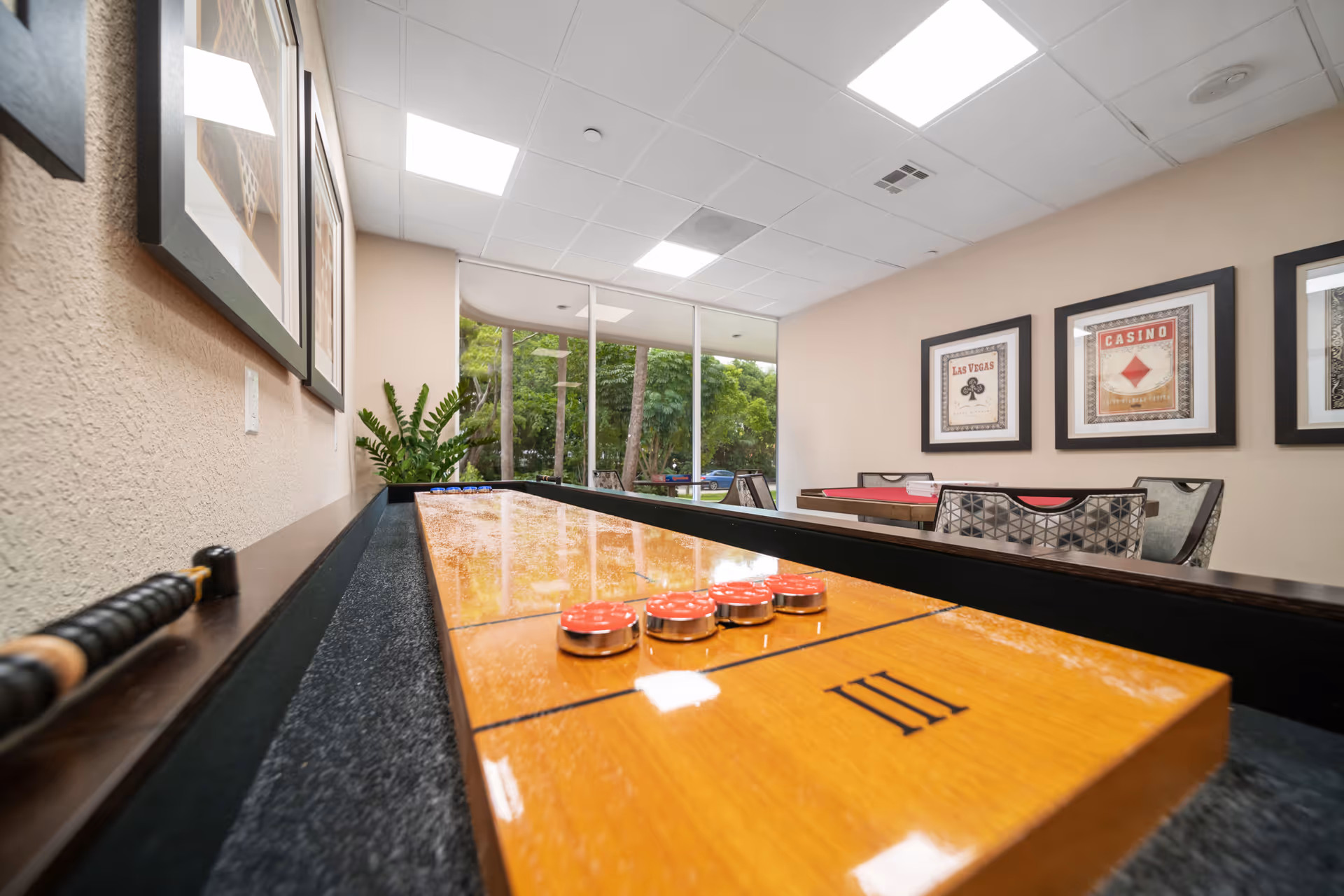Interior common room featuring a shuffleboard table with game pucks in the foreground, seating, casino-themed framed art, and large windows overlooking trees.