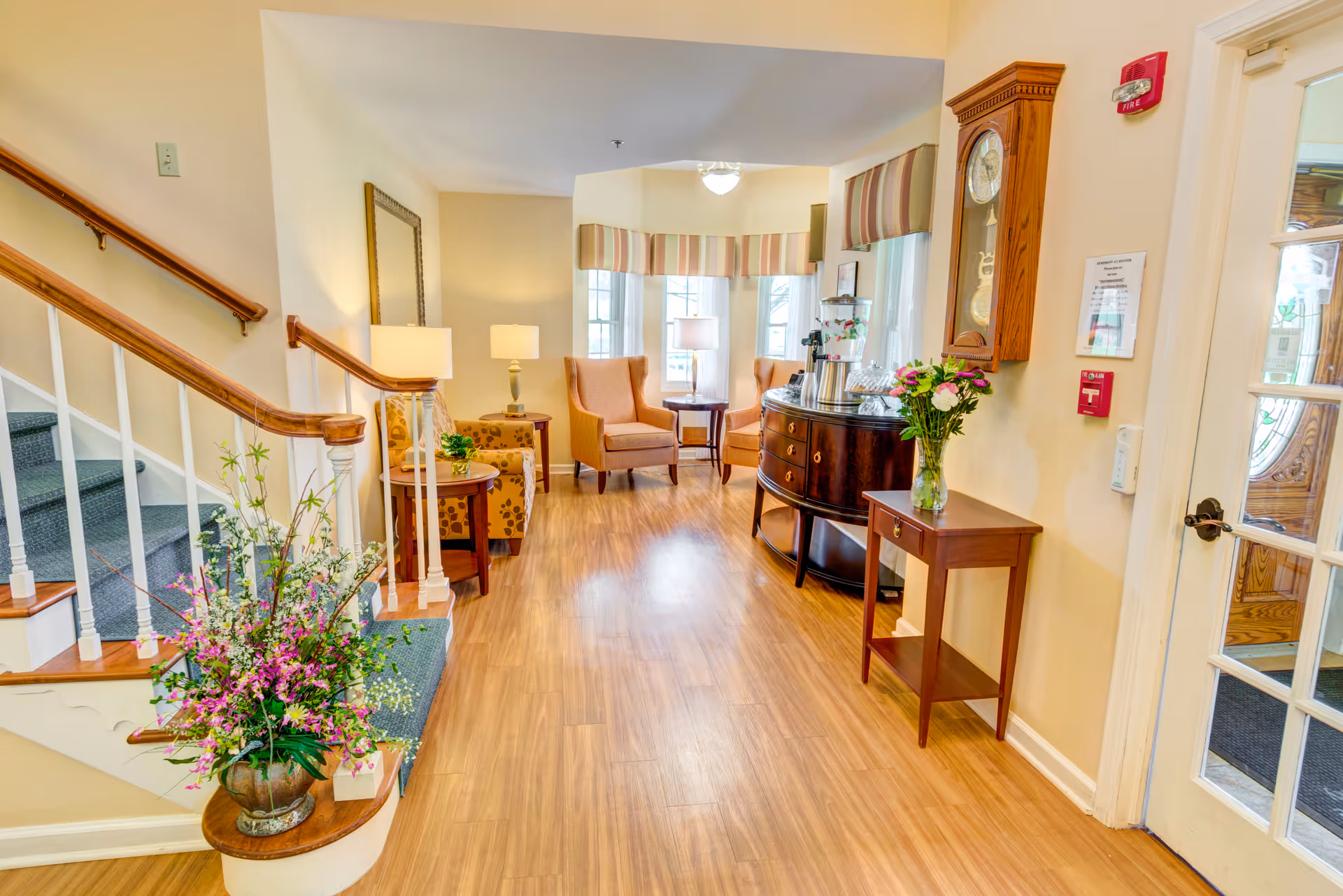 A bright and welcoming interior common area with wooden flooring, a staircase with wooden handrails on the left, a large floral arrangement on a small round table near the stairs, two upholstered armchairs with a small table and lamp between them near the back wall, a wooden sideboard with a water dispenser and flowers on top, a wall clock, and a glass door on the right side.