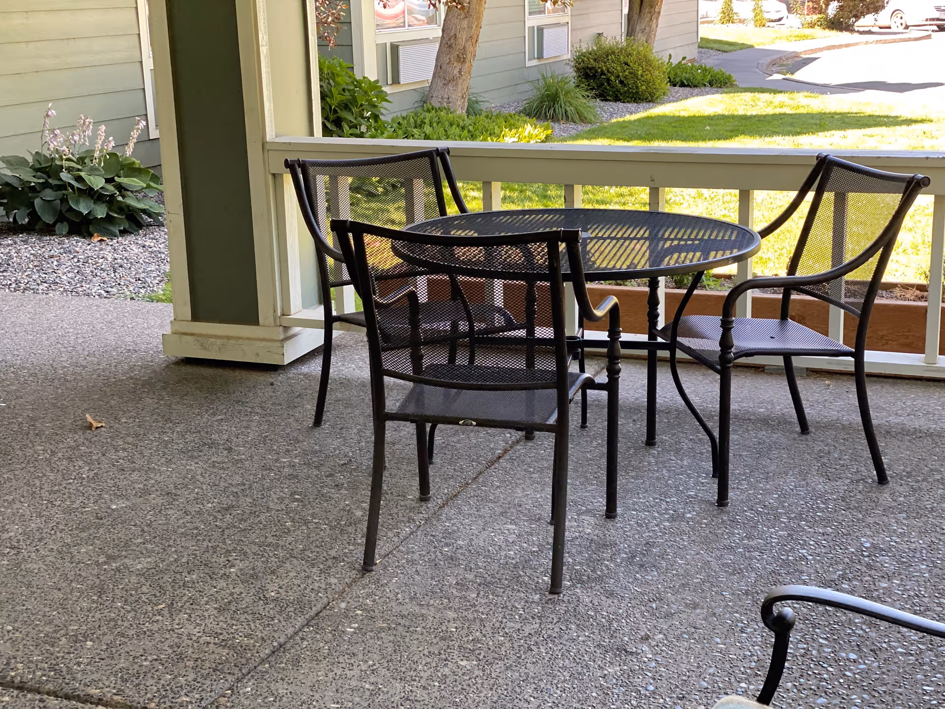 Outdoor patio area with a round metal table and four matching metal chairs on a concrete floor. The patio is partially enclosed with a railing and overlooks a grassy area with trees and shrubs.