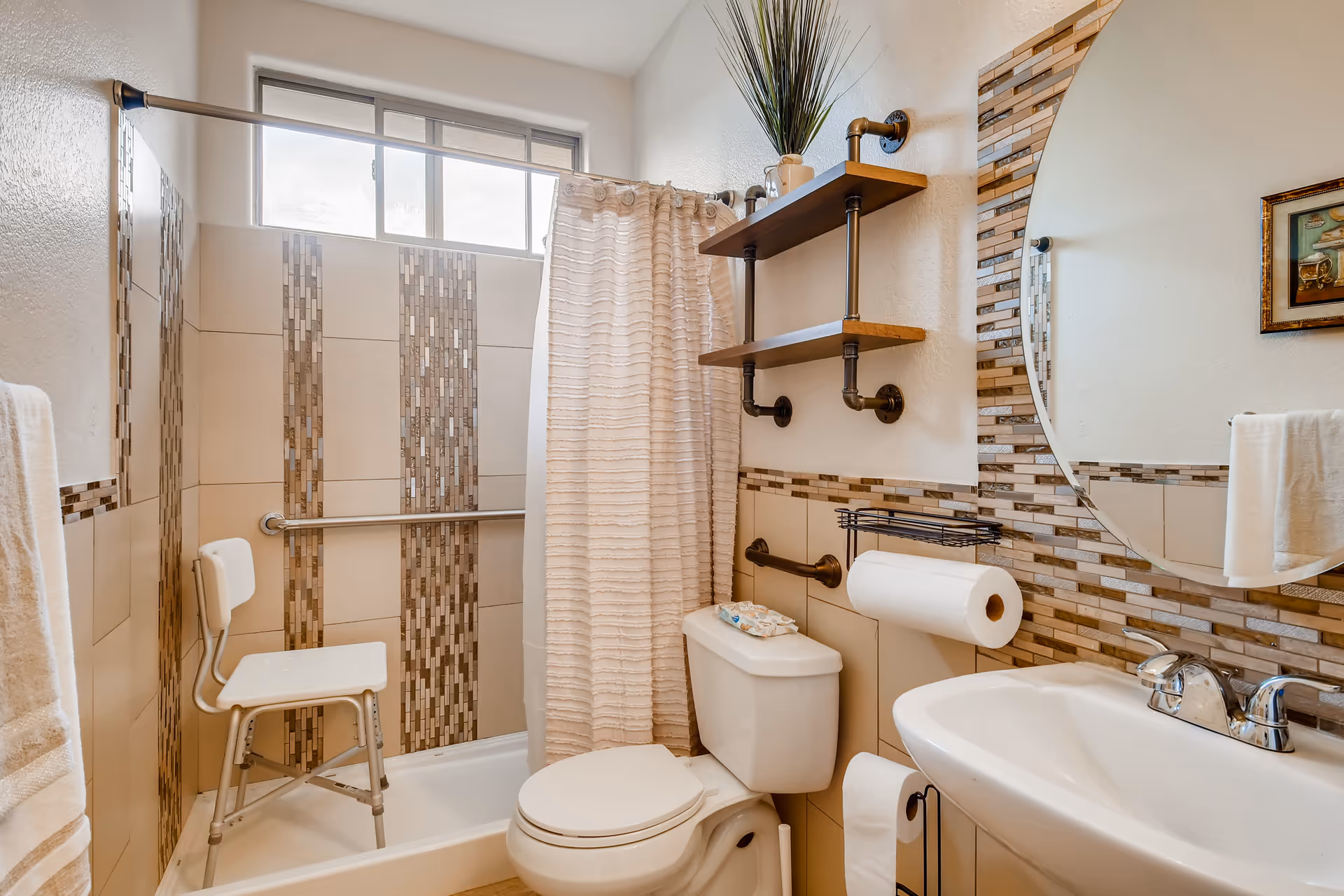 A bathroom with a white toilet, a white sink with a chrome faucet, and a shower area with a white shower chair and a beige shower curtain. The walls have beige tiles with vertical mosaic tile accents. There are two wooden shelves with a potted plant above the toilet, a round mirror with mosaic tile border, and a towel hanging on a rack.