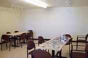A plain room with white walls and ceiling, containing two rectangular tables with several chairs around them. The tables have some items on them, possibly puzzles or games, and the room is lit by fluorescent ceiling lights.