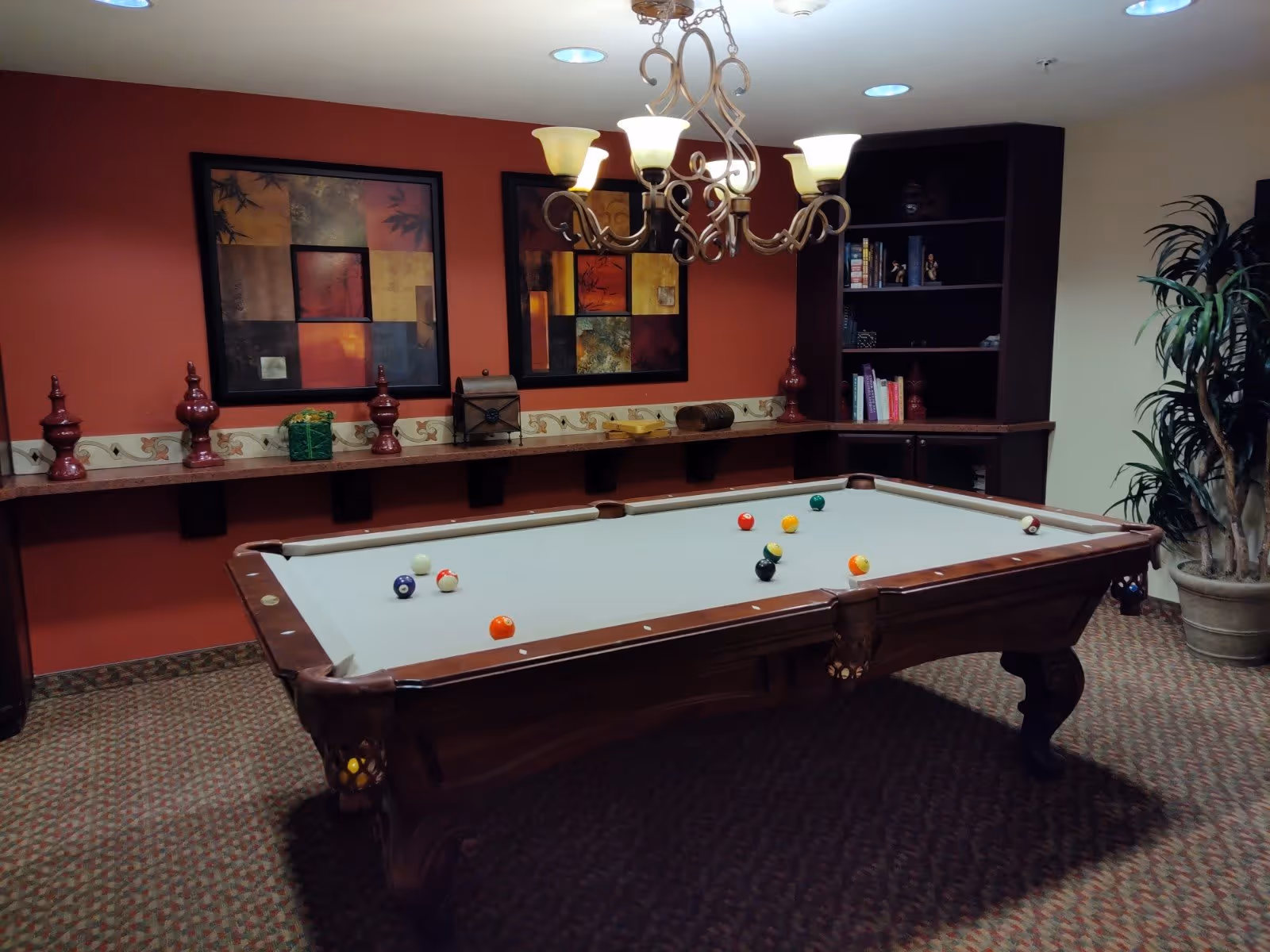 A room with a pool table in the center, several billiard balls scattered on the table. The walls are painted red with two abstract paintings hanging. A chandelier with multiple lights hangs above the pool table. There is a long shelf with decorative items and a dark wooden bookshelf filled with books and small figurines. A large potted plant is in the corner of the room.