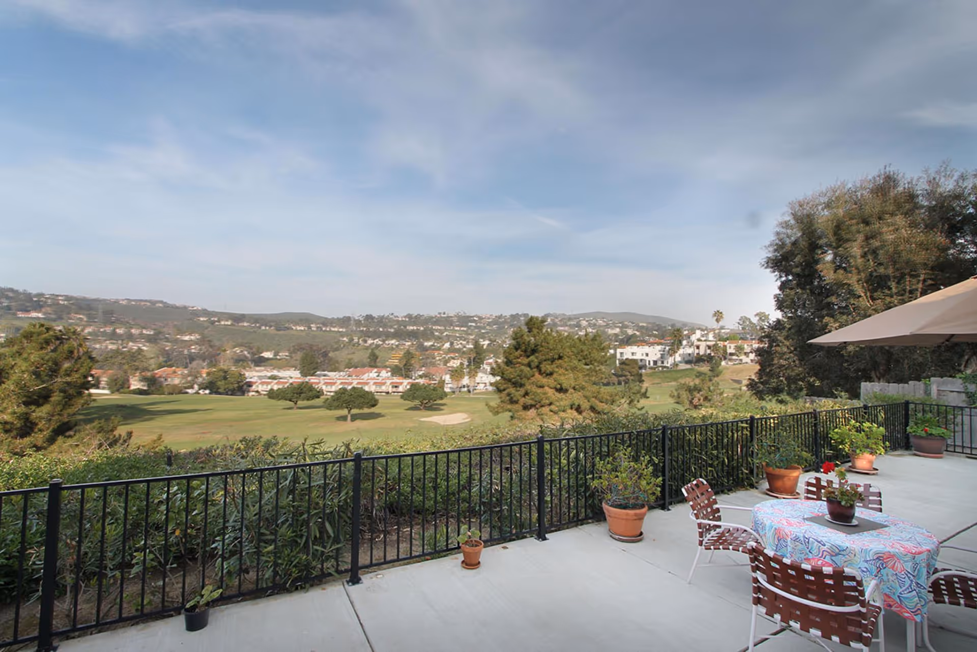 Patio with a table, chairs, potted plants and an umbrella overlooking a golf course and distant hills.