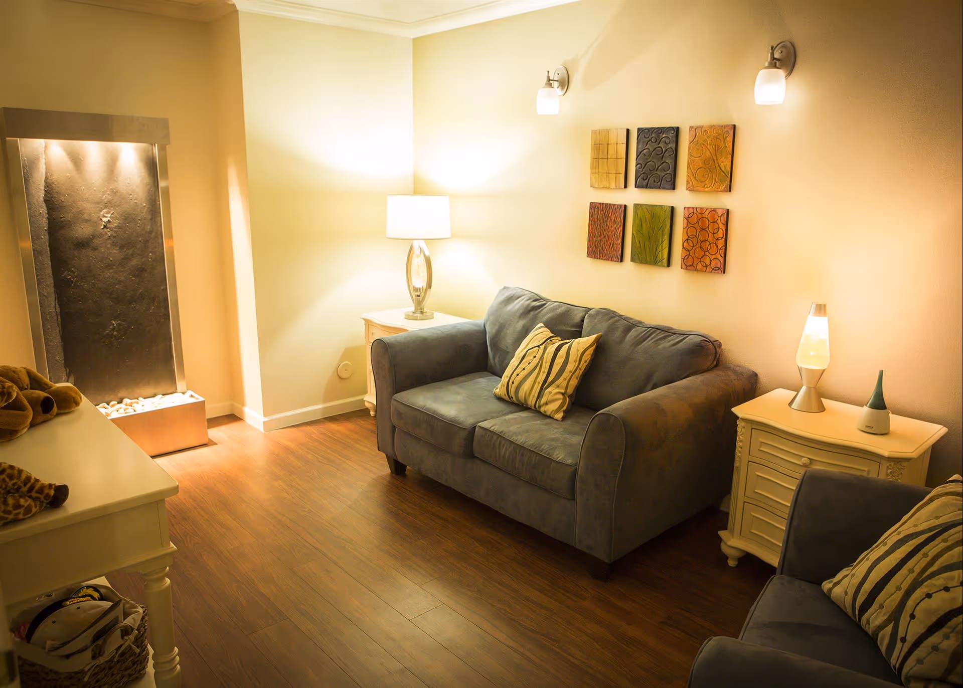 Cozy living room with two gray upholstered sofas, each with a striped pillow. The room features warm lighting from a table lamp on a white side table and a lava lamp on another white side table. There is a decorative water feature with stones in the corner and six colorful square wall art pieces arranged in two rows above one sofa. The floor is wooden, and the walls are painted light beige.