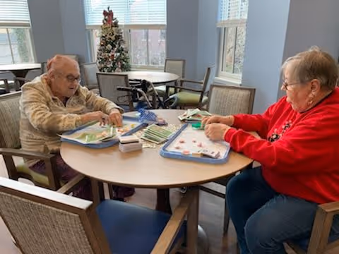 Two elderly women sitting at a round table in a well-lit room, playing a board game. There is a decorated Christmas tree in the background near the windows.