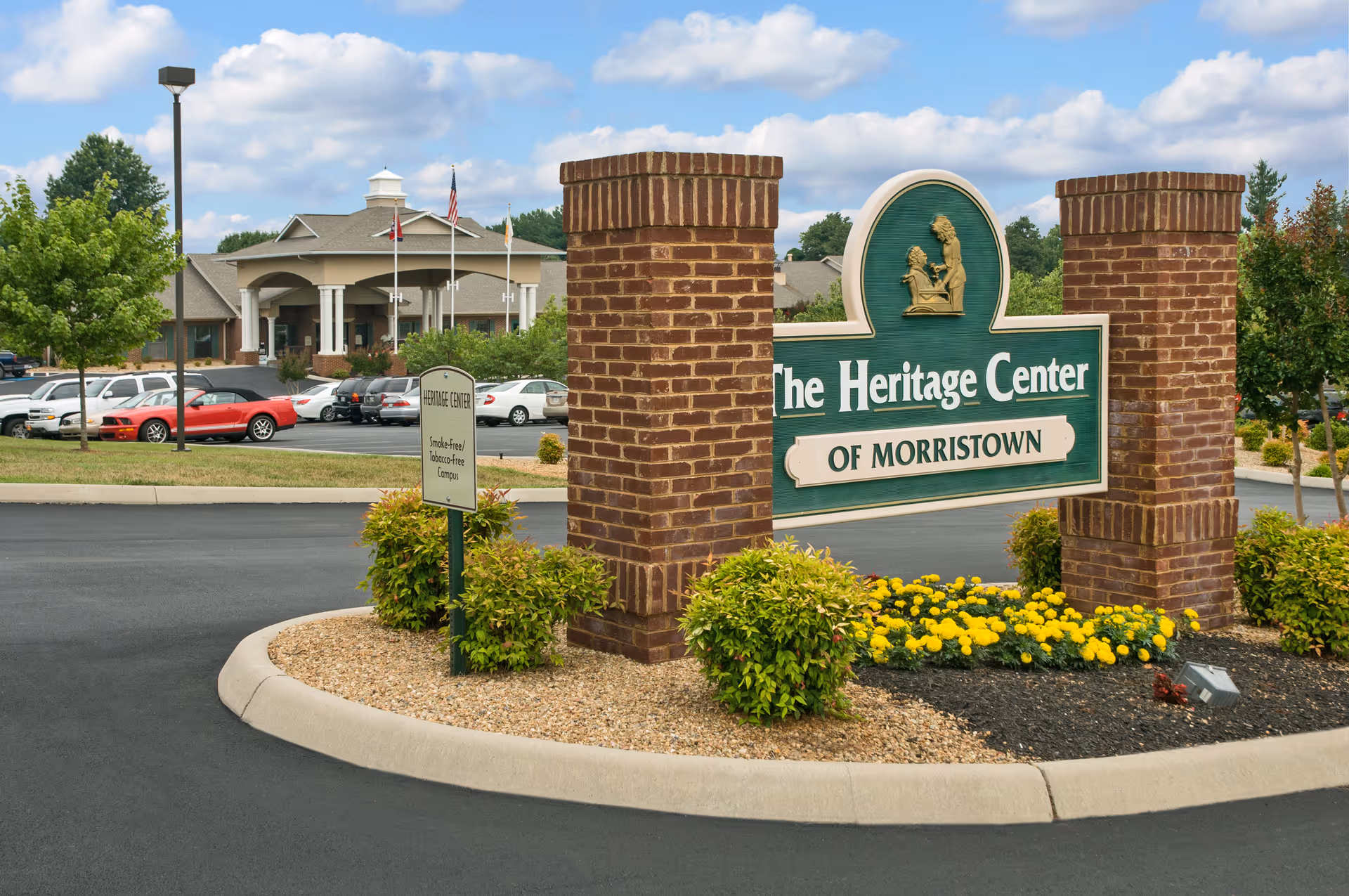 Large brick entrance sign reading "The Heritage Center of Morristown" in front of the facility building, parking lot, and landscaping.
