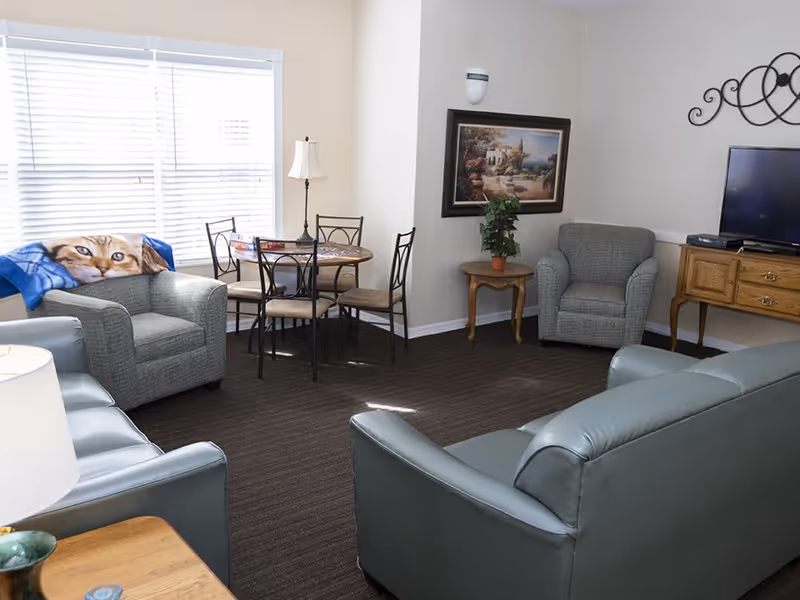 A bright common living room with sofas, armchairs, a small round dining table, and a TV on a wooden cabinet.