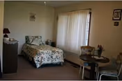 Small bedroom with a single bed in floral bedding, a dresser, a round table with chairs, and a large curtained window.