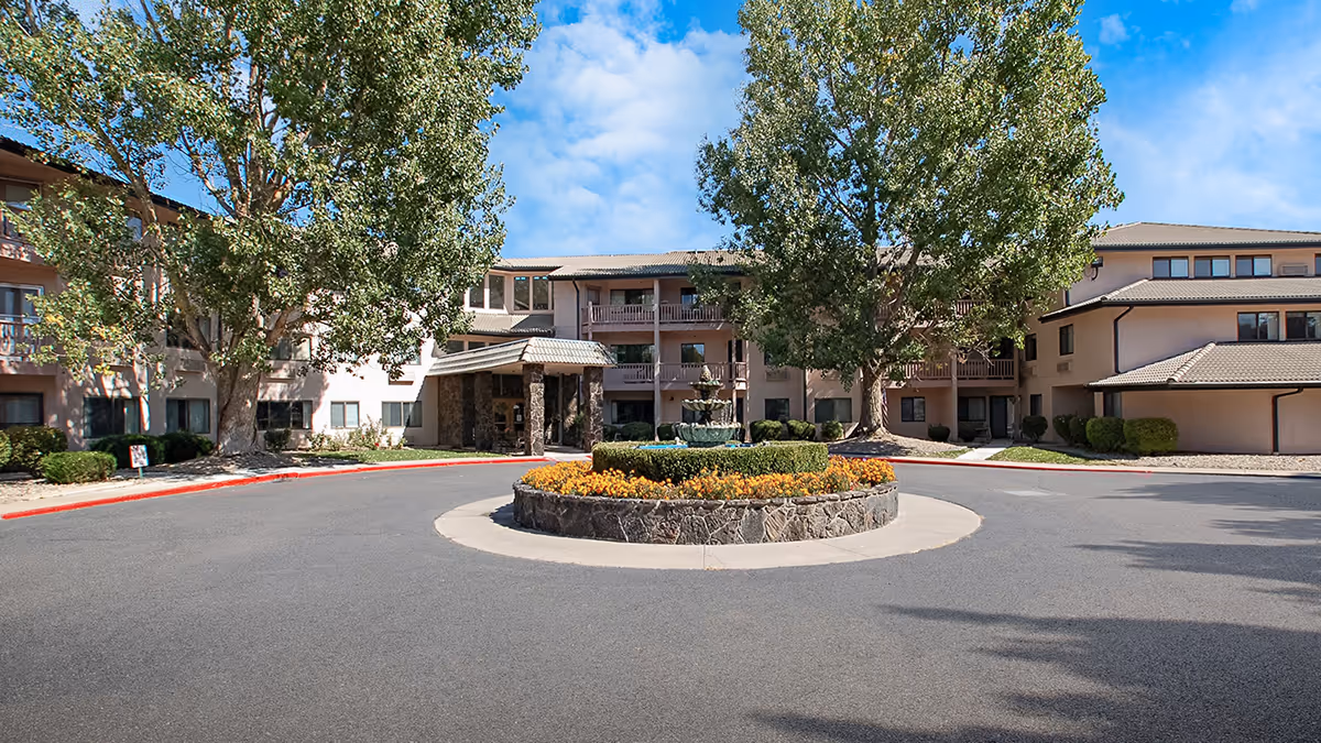 Front exterior of a multi-story senior living building with a circular driveway, central flowerbed and fountain, and mature trees.