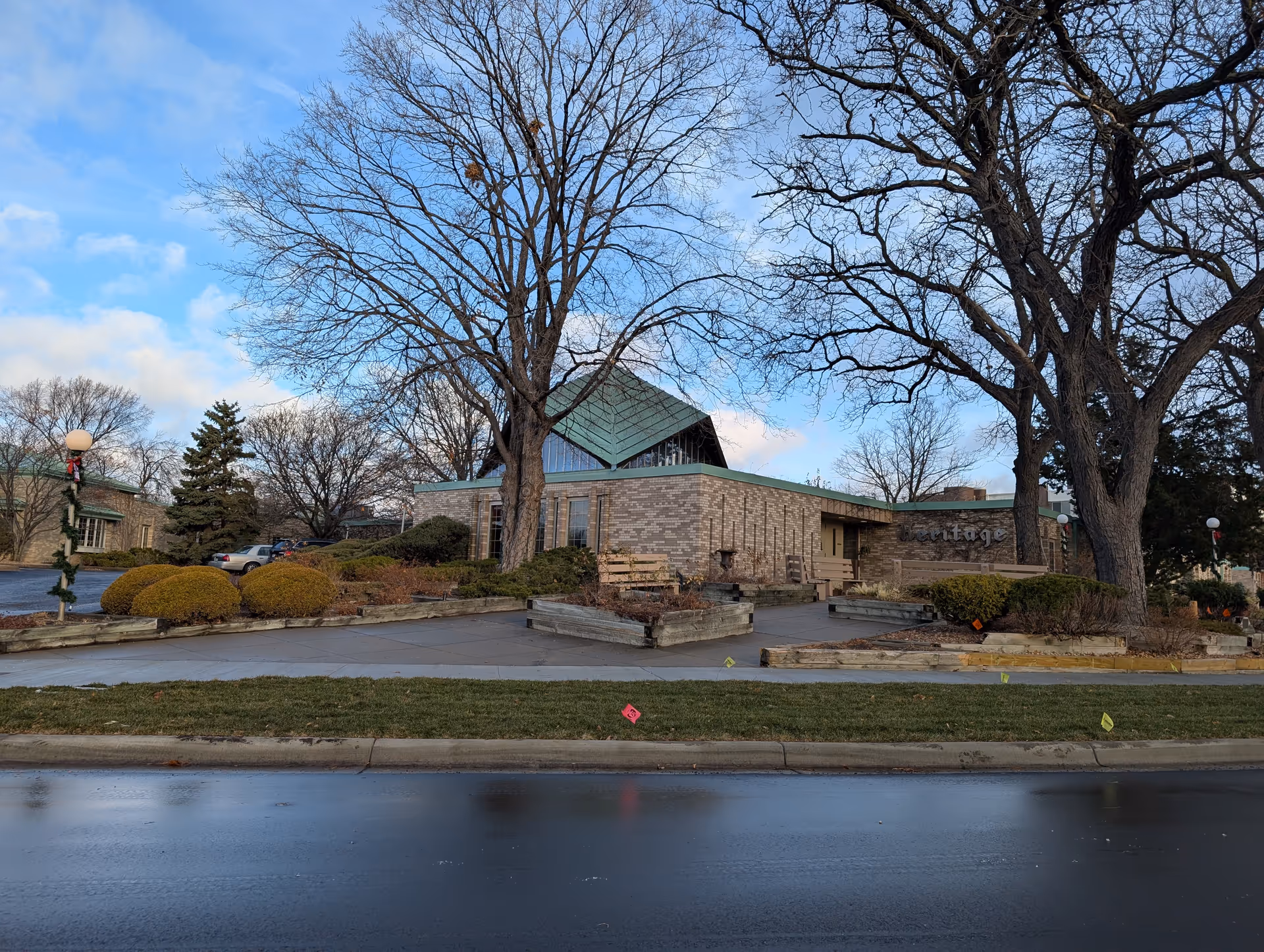 Exterior view of a single-story brick building with a green roof, surrounded by leafless trees and landscaped bushes. The building has benches and a sign partially visible that reads 'Heritage'. The sky is partly cloudy and the street in front is wet.