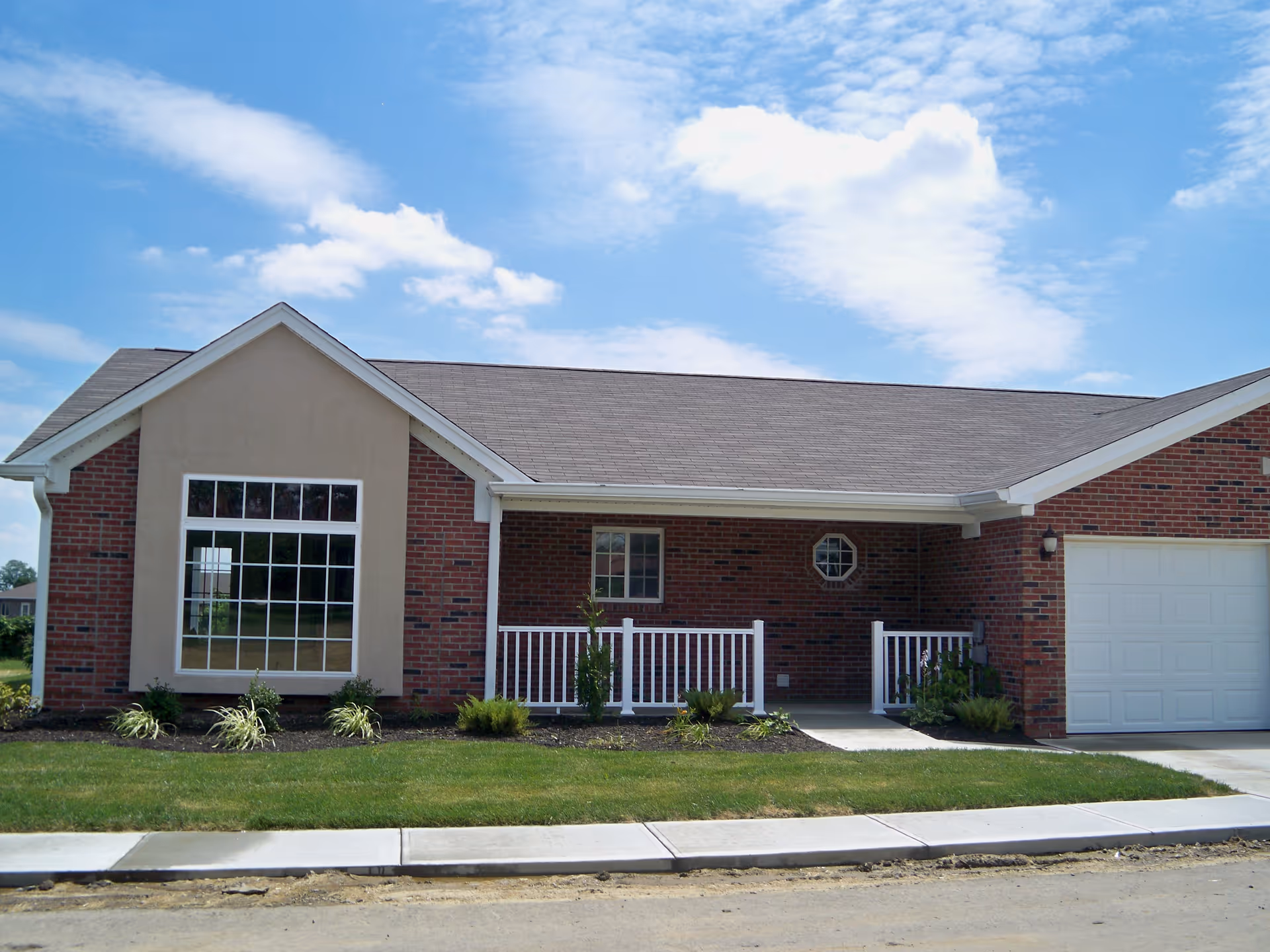 Front exterior view of a single-story brick and stucco building with a large window, white railing porch, garage door, and a well-maintained lawn under a partly cloudy blue sky.