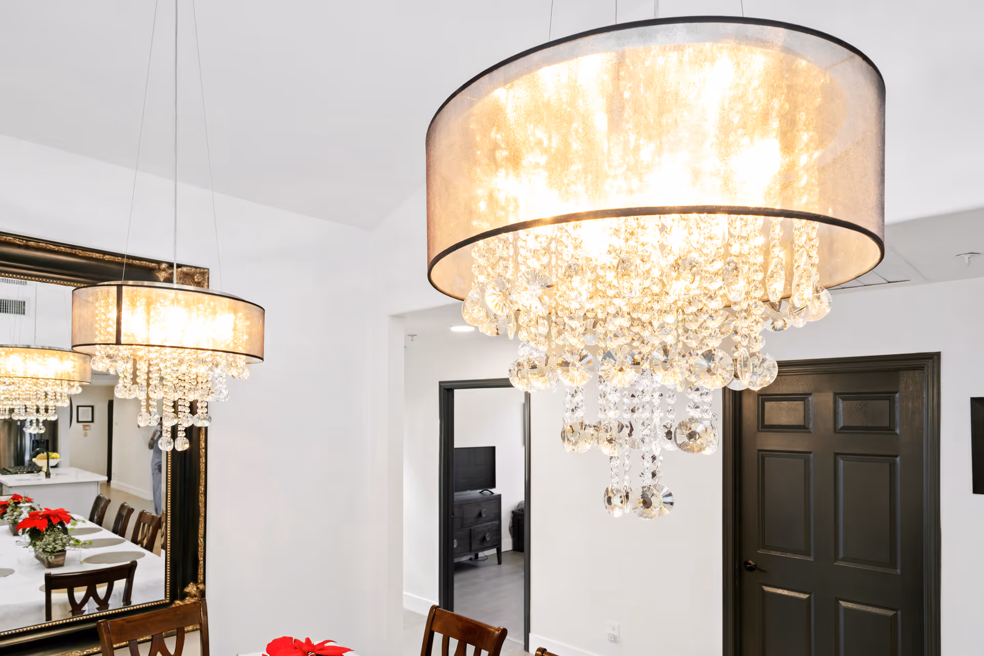 Interior view of a dining area with a large, elegant chandelier featuring hanging crystal ornaments. A large mirror on the wall reflects the chandelier and a dining table set with chairs and a white tablecloth with red floral centerpieces. A dark door and an open doorway leading to another room with a TV and furniture are visible.