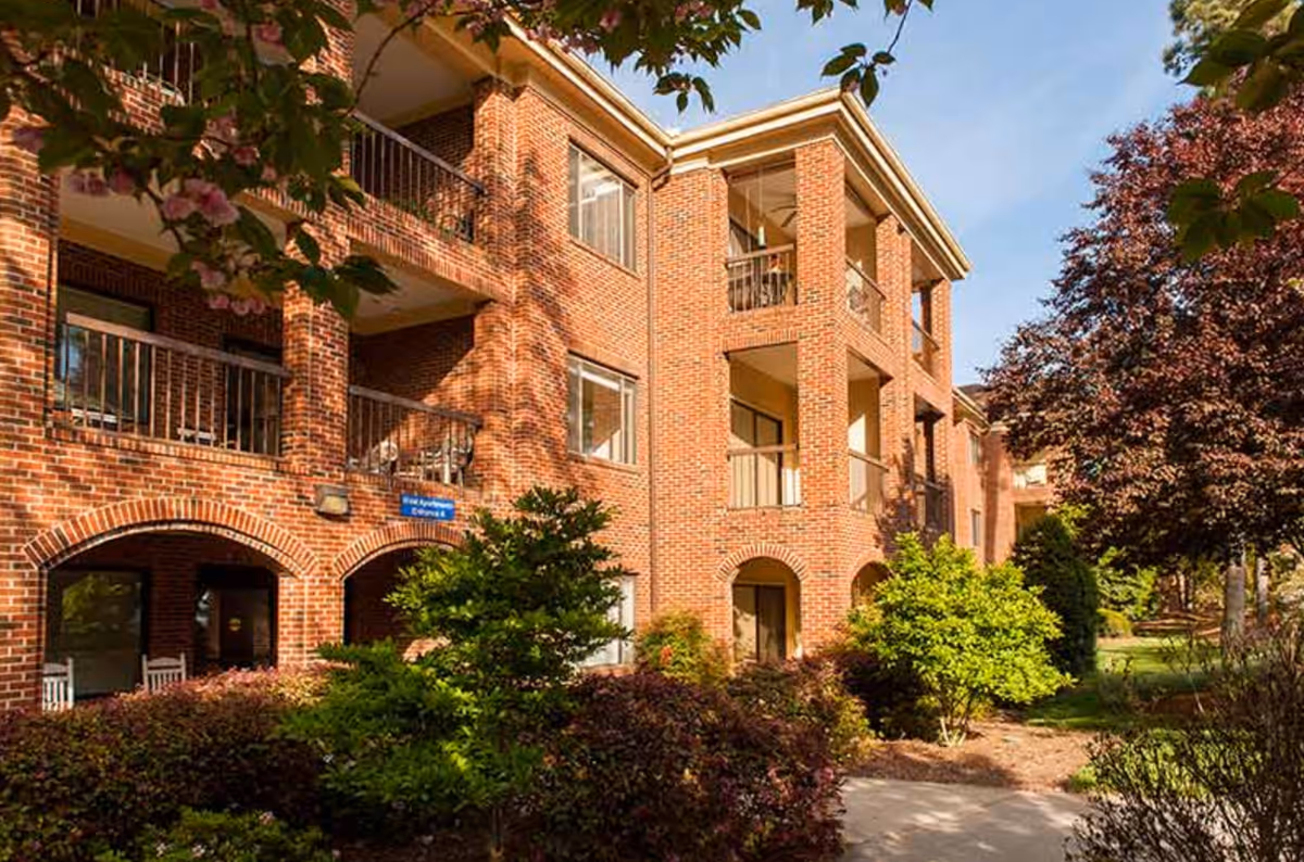 Three-story red brick retirement community building with balconies and landscaped front gardens.