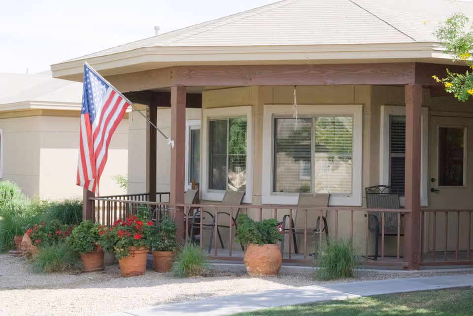 Front porch area of a senior living facility with several chairs, potted plants, and an American flag mounted on a wooden post. The porch is covered and has windows and a door leading inside.