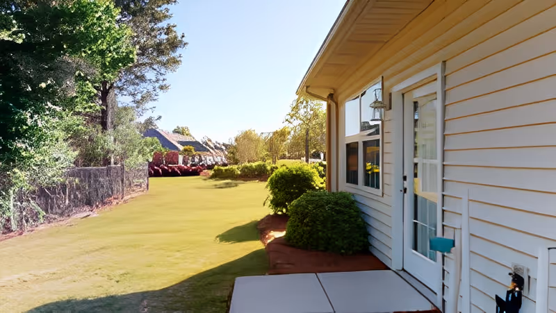 Side exterior of a light-colored cottage with a small concrete patio and door opening onto a grassy yard with shrubs and trees.