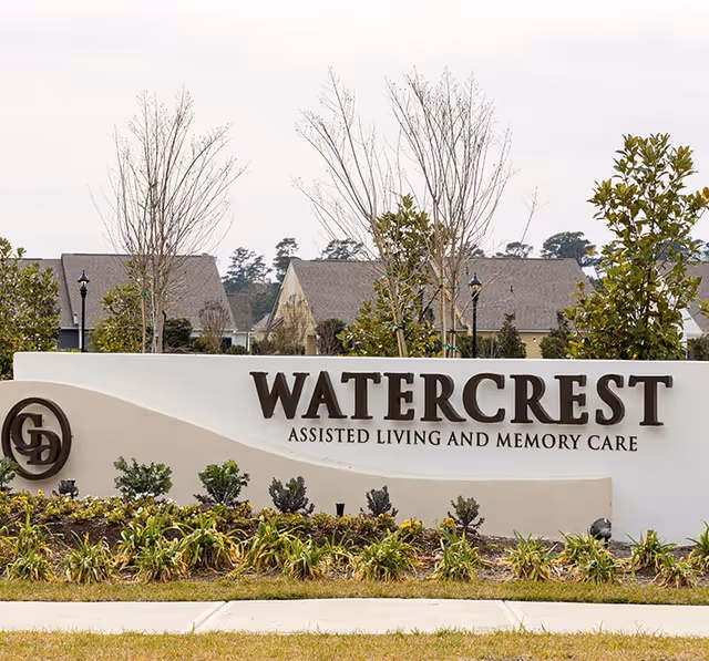 Entrance sign reading "WATERCREST Assisted Living and Memory Care" with landscaping and homes behind it.